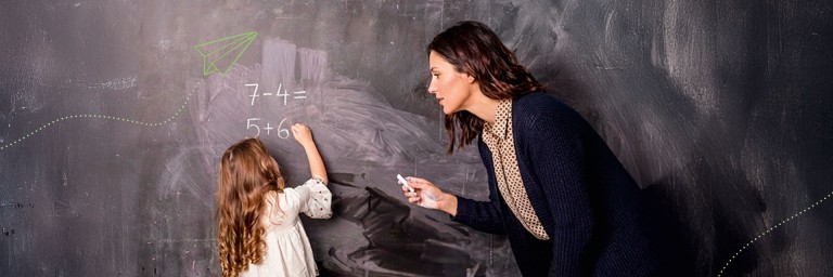 Ein kleines Kind steht an der Tafel und schreibt eine Rechenaufgabe mit Kreide auf. Daneben eine Lehrerin und hilft.