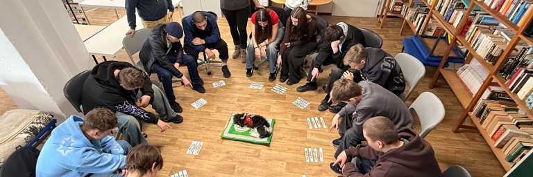 Gruppe von jungen Leuten sitzen im Stuhlkreis zusammen und in der Mitte liegt ein Hund.
