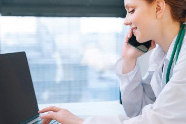 Eine junge Frau mit weißem Kittel am Laptop und telefoniert dabei.