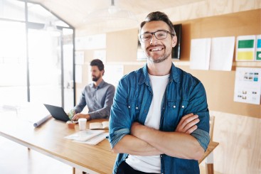 Mann lächelt mit verschränkten Armen in die Kamera, im Hintergrund sitzt ein Mann an einem Holz-Schreibtisch