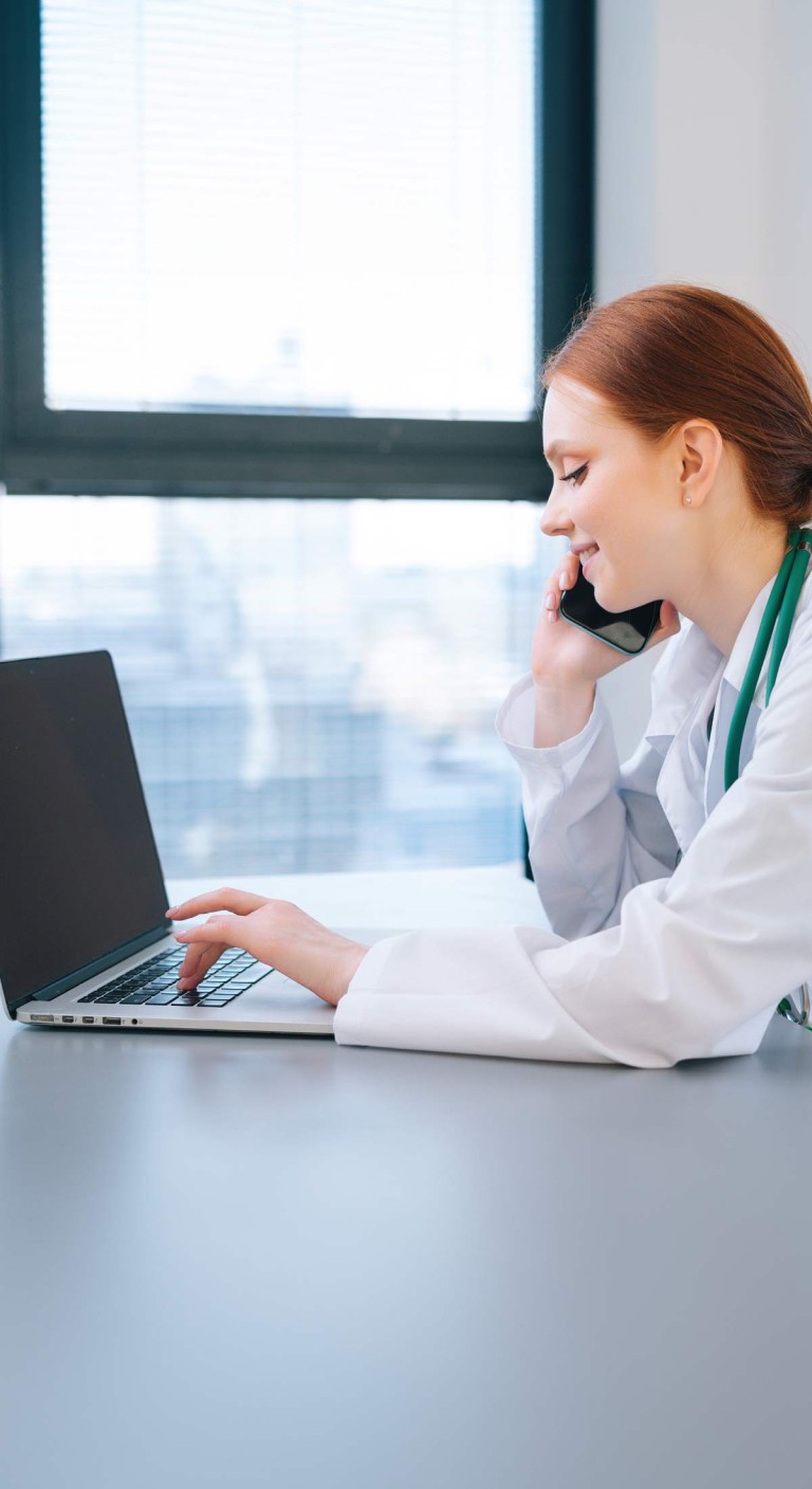Eine junge Frau mit weißem Kittel am Laptop und telefoniert dabei.