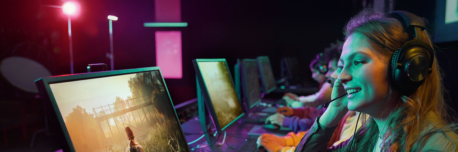 Junge Frau mit Kopfhörern sitzt im dunklen vor einem Monitor. Neben ihr weitere Menschen vorn Monitoren.