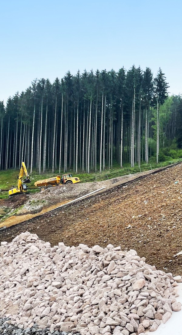 Ein Bagger und ein LKW stehen auf einem kahlen Hang am Wald. Im Vordergrund liegt ein großer Haufen Steine.